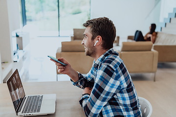 Image showing The man sitting at a table in a modern living room, with headphones using a laptop and smartphone for business video chat, conversation with friends and entertainment