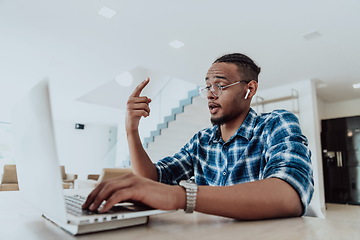 Image showing African American man in glasses sitting at a table in a modern living room, using a laptop for business video chat, conversation with friends and entertainment