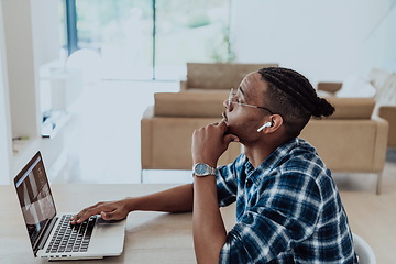 Image showing African American man in glasses sitting at a table in a modern living room, using a laptop for business video chat, conversation with friends and entertainment