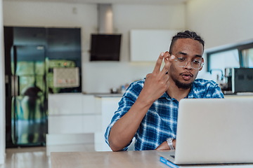 Image showing African American man in glasses sitting at a table in a modern living room, using a laptop for business video chat, conversation with friends and entertainment