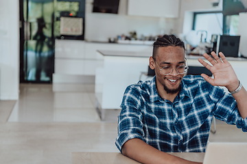 Image showing African American man in glasses sitting at a table in a modern living room, using a laptop for business video chat, conversation with friends and entertainment