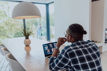 Image showing African American man in glasses sitting at a table in a modern living room, using a laptop and smartphone for business video chat, conversation with friends and entertainment