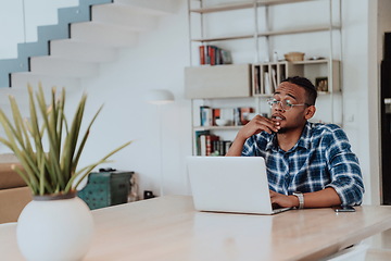 Image showing African American man in glasses sitting at a table in a modern living room, using a laptop for business video chat, conversation with friends and entertainment