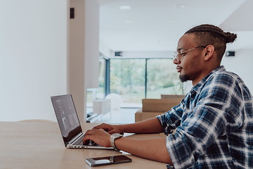 Image showing African American man in glasses sitting at a table in a modern living room, using a laptop for business video chat, conversation with friends and entertainment