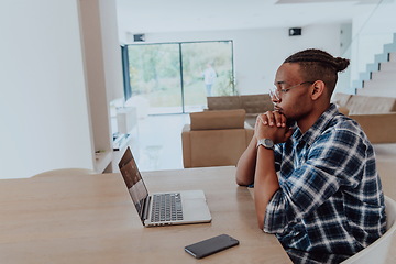 Image showing African American man in glasses sitting at a table in a modern living room, using a laptop for business video chat, conversation with friends and entertainment