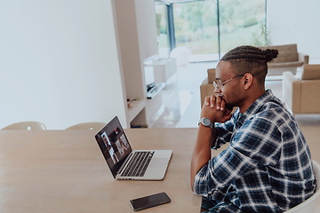 Image showing African American man in glasses sitting at a table in a modern living room, using a laptop for business video chat, conversation with friends and entertainment