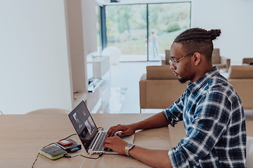 Image showing African American man in glasses sitting at a table in a modern living room, using a laptop for business video chat, conversation with friends and entertainment