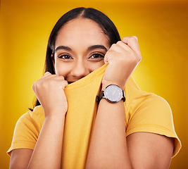 Image showing Portrait, shy and funny with a woman on a yellow background in studio pulling on her t-shirt. Face, fashion or playful and an attractive young female covering her mouth with trendy clothes for style