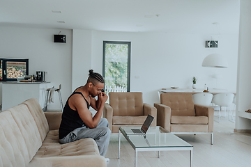 Image showing African American sport man in glasses sitting at a table in a modern living room, using a laptop for business video chat, conversation with friends and entertainment