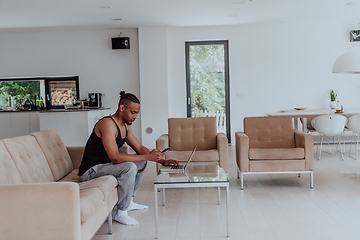 Image showing African American sport man in glasses sitting at a table in a modern living room, using a laptop for business video chat, conversation with friends and entertainment