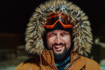 Image showing Head shot of a man in a cold snowy area wearing a thick brown winter jacket, snow goggles and gloves on a cold Scandinavian night. Life in the cold regions of the country.