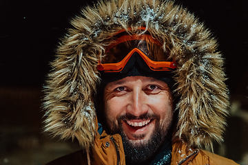 Image showing Head shot of a man in a cold snowy area wearing a thick brown winter jacket, snow goggles and gloves on a cold Scandinavian night. Life in the cold regions of the country.