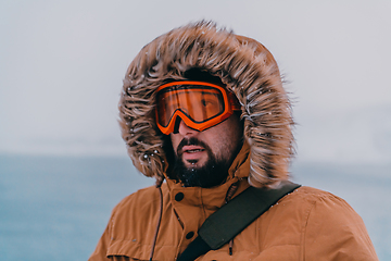 Image showing Headshot photo of a man in a cold snowy area wearing a thick brown winter jacket, snow goggles and gloves. Life in cold regions of the country.