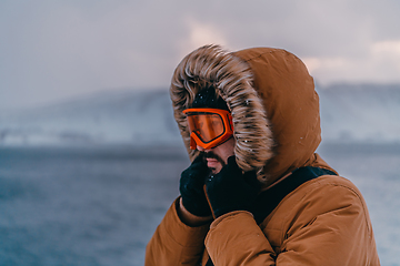 Image showing Headshot photo of a man in a cold snowy area wearing a thick brown winter jacket, snow goggles and gloves. Life in cold regions of the country.