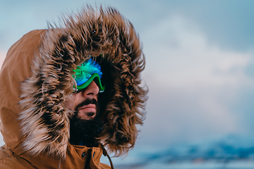 Image showing Headshot photo of a man in a cold snowy area wearing a thick brown winter jacket, snow goggles and gloves. Life in cold regions of the country.