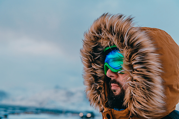 Image showing Headshot photo of a man in a cold snowy area wearing a thick brown winter jacket, snow goggles and gloves. Life in cold regions of the country.
