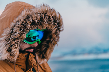Image showing Headshot photo of a man in a cold snowy area wearing a thick brown winter jacket, snow goggles and gloves. Life in cold regions of the country.