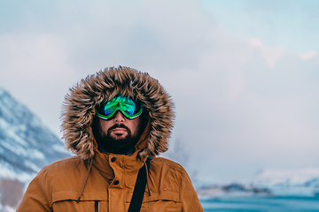 Image showing Headshot photo of a man in a cold snowy area wearing a thick brown winter jacket, snow goggles and gloves. Life in cold regions of the country.