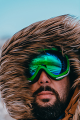 Image showing Headshot photo of a man in a cold snowy area wearing a thick brown winter jacket, snow goggles and gloves. Life in cold regions of the country.