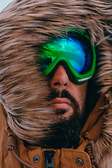 Image showing Headshot photo of a man in a cold snowy area wearing a thick brown winter jacket, snow goggles and gloves. Life in cold regions of the country.