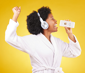 Image showing Black woman, music headphones and singing with phone in studio isolated on a yellow background. Karaoke singer, bathrobe and happy female dance with mobile microphone while streaming podcast or radio