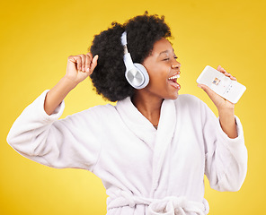 Image showing Black woman, singing and happiness in a bathrobe with phone in studio isolated on a yellow background. Singer, headphones or happy female dance with mobile microphone while streaming podcast or radio
