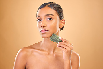 Image showing Face portrait, skincare and woman with gua sha in studio isolated on a brown background. Dermatology, massage and serious Indian female model with jade crystal or stone for healthy skin treatment.