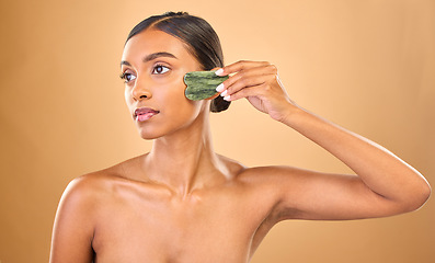 Image showing Face, beauty skincare and woman with gua sha in studio isolated on a brown background. Dermatology, thinking and serious Indian female model with jade crystal or stone for healthy skin treatment.