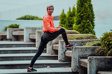 Image showing Fit attractive woman in sportswear stretching one leg before jogging on the footpath outdoor in summer among greenery. Workout, sport, activity, fitness, vacation and training concept.