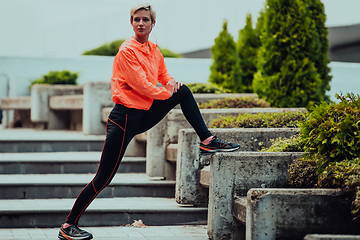 Image showing Fit attractive woman in sportswear stretching one leg before jogging on the footpath outdoor in summer among greenery. Workout, sport, activity, fitness, vacation and training concept.