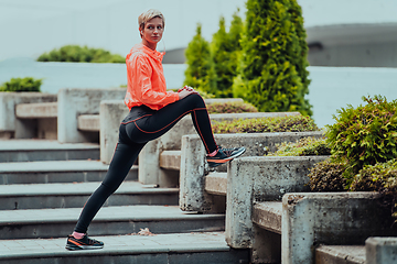 Image showing Fit attractive woman in sportswear stretching one leg before jogging on the footpath outdoor in summer among greenery. Workout, sport, activity, fitness, vacation and training concept.