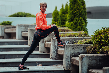 Image showing Fit attractive woman in sportswear stretching one leg before jogging on the footpath outdoor in summer among greenery. Workout, sport, activity, fitness, vacation and training concept.