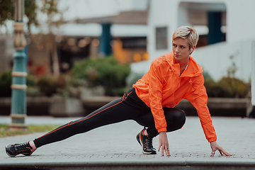 Image showing Fit attractive woman in sportswear stretching one leg before jogging on the footpath outdoor in summer among greenery. Workout, sport, activity, fitness, vacation and training concept.