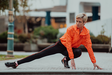 Image showing Fit attractive woman in sportswear stretching one leg before jogging on the footpath outdoor in summer among greenery. Workout, sport, activity, fitness, vacation and training concept.