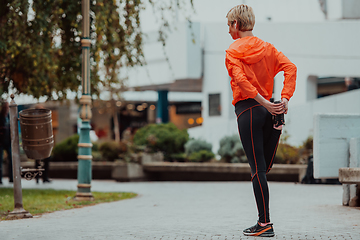 Image showing Fit attractive woman in sportswear stretching one leg before jogging on the footpath outdoor in summer among greenery. Workout, sport, activity, fitness, vacation and training concept.