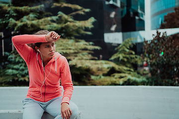 Image showing a woman in a sports outfit is resting in a city environment after a hard morning workout while using noiseless headphones