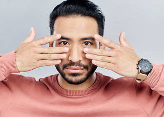 Image showing Portrait, eyes and vision with a man in studio on a gray background making a masquerade hand gesture. Face, hands and focus with a handsome young male posing against a wall while touching his cheeks