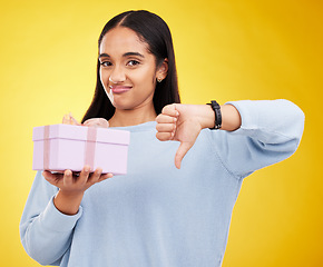 Image showing Disappointed, portrait and woman with a thumbs down for a gift isolated on a yellow background. Unhappy, negative and a girl holding a present and showing a hand gesture for disappointment in studio