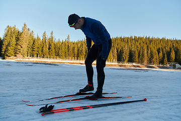 Image showing Handsome male athlete with cross country skis preparing equipment for training in a snowy forest. Checking smartwatch. Healthy winter lifestyle.