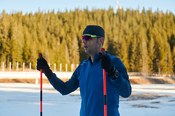 Image showing Handsome male athlete with cross country skis preparing equipment for training in a snowy forest. Checking smartwatch. Healthy winter lifestyle.