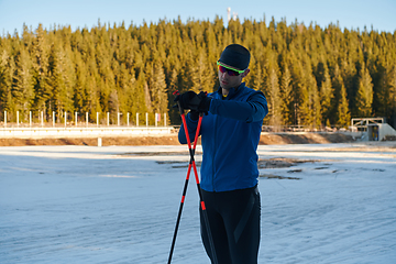 Image showing Handsome male athlete with cross country skis, taking fresh breath and having break after hard workout training in a snowy forest. Checking smartwatch. Healthy winter lifestyle