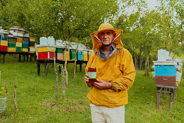 Image showing The beekeeper holding a jar of honey in his hand while standing in a meadow surrounded by a box and a honey farm