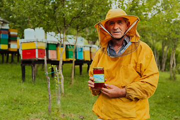 Image showing The beekeeper holding a jar of honey in his hand while standing in a meadow surrounded by a box and a honey farm