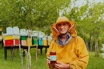 Image showing The beekeeper holding a jar of honey in his hand while standing in a meadow surrounded by a box and a honey farm