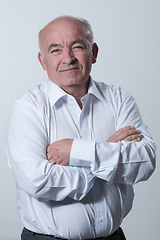 Image showing Confident senior man in white shirt crossing hands on chest and looking at camera while standing against gray background. Self confident senior isolated white studio shoot.