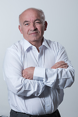 Image showing Confident senior man in white shirt crossing hands on chest and looking at camera while standing against gray background. Self confident senior isolated white studio shoot.