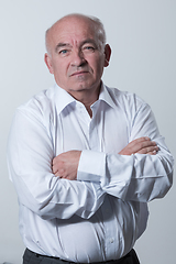 Image showing Confident senior man in white shirt crossing hands on chest and looking at camera while standing against gray background. Self confident senior isolated white studio shoot.