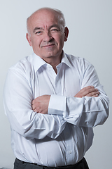 Image showing Confident senior man in white shirt crossing hands on chest and looking at camera while standing against gray background. Self confident senior isolated white studio shoot.