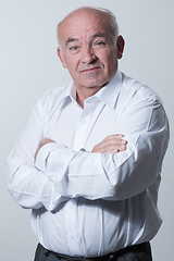 Image showing Confident senior man in white shirt crossing hands on chest and looking at camera while standing against gray background. Self confident senior isolated white studio shoot.