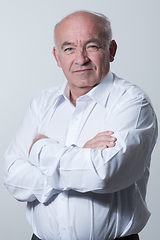 Image showing Confident senior man in white shirt crossing hands on chest and looking at camera while standing against gray background. Self confident senior isolated white studio shoot.